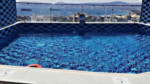 A rooftop pool with a checkered tile border, clear blue water, and a view of a cityscape and the sea. - Alena Hotel (Aqaba)