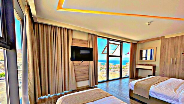 A hotel room with two beds, a TV, a large window with a view of the beach, and a wooden floor. - Alena Hotel (Aqaba)