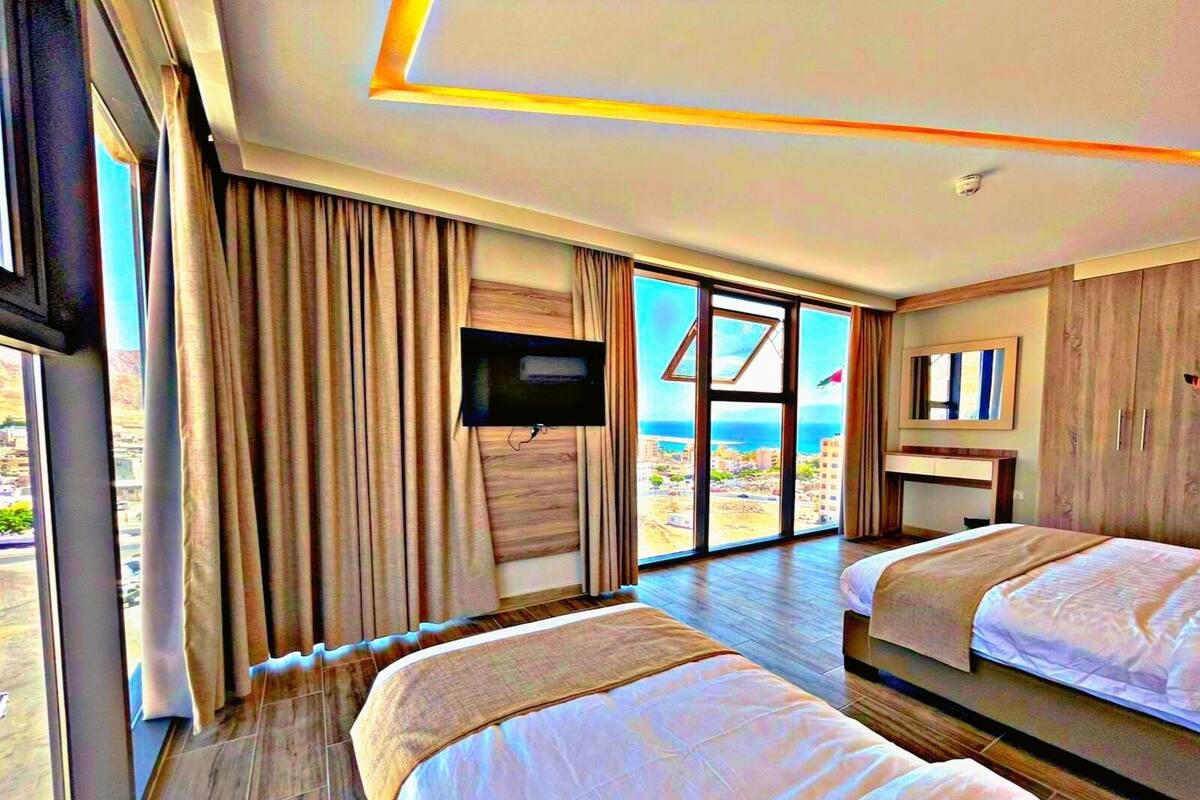 A hotel room with two beds, a TV, a large window with a view of the beach, and a wooden floor.