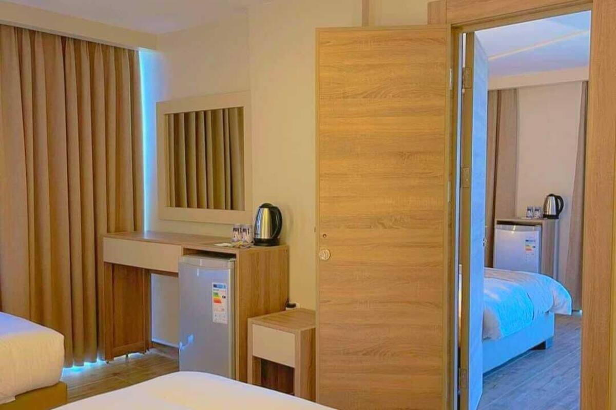 A hotel room with a bed, a desk with a kettle, a minibar, and a view of another room.