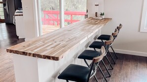 Una cocina moderna con una isla de madera y taburetes de bar, ventanas amplias y una terraza roja visible a través del vidrio.