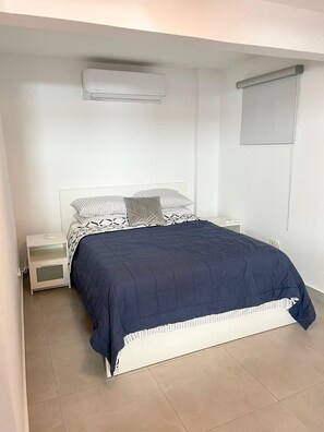 A bedroom with a bed, a nightstand, a wall-mounted air conditioner, and a window with a blind.