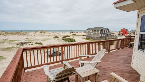 Eine Holzdar Terrasse mit zwei Holzstühlen und einem Tisch, mit Blick auf einen Strand mit Dünen und Häusern im Hintergrund.