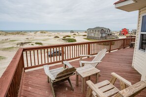 A wooden deck with two wooden chairs and a table, overlooking a beach with sand dunes and houses in the background. - Corolla 4x4 beach cottage with phenomenal ocean views! (Corolla)