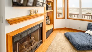 Ein gemütliches Wohnzimmer mit Kamin, Holzregalen und Blick auf den Strand durch große Fenster.