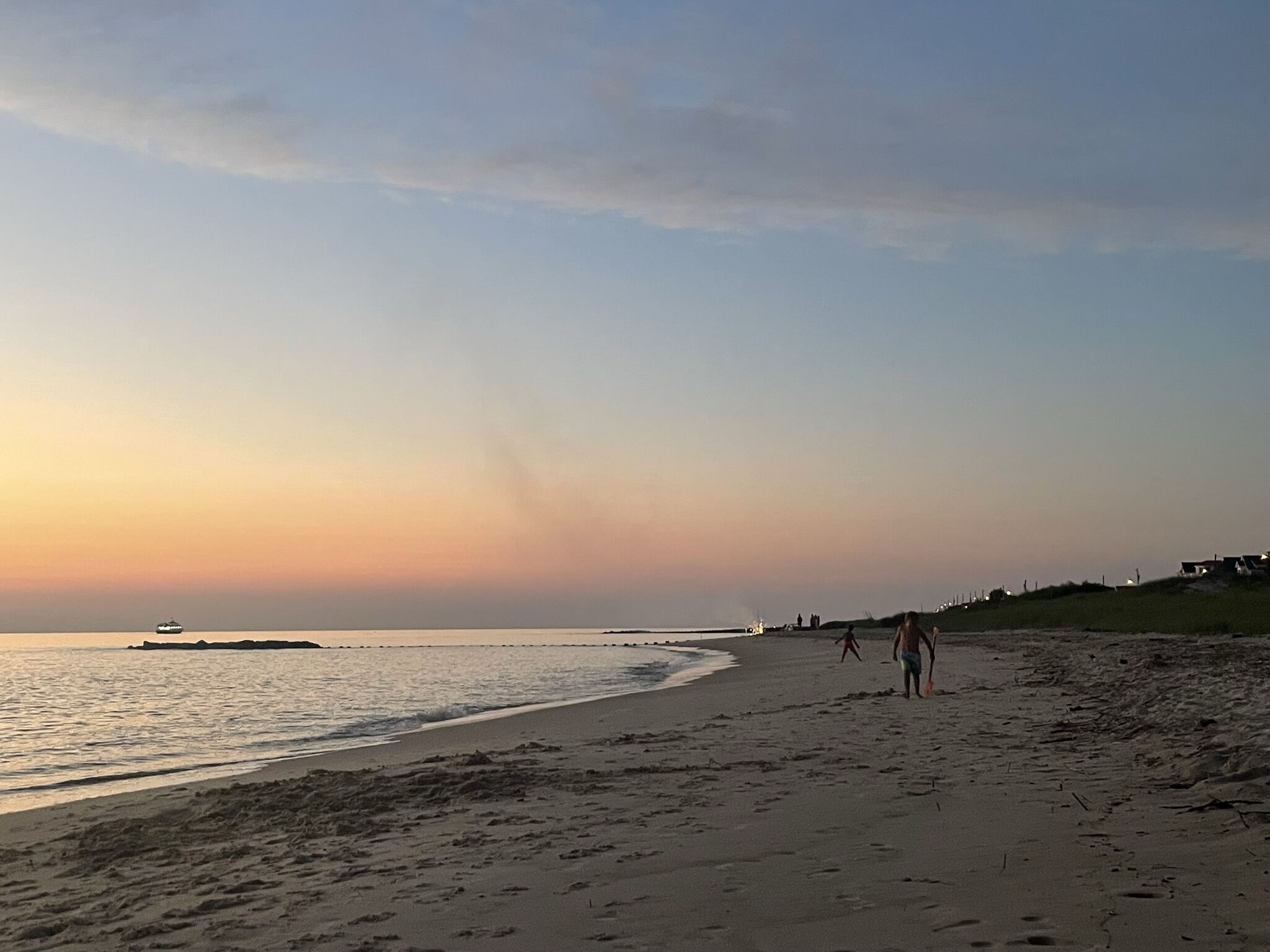 Een strand met een rustige zee, enkele mensen die wandelen en een boot in de verte.