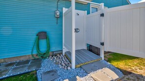 Un espace extérieur clos, agrémenté d’une clôture, d’un portail en bois, d’un tuyau d’arrosage et de dalles qui mènent à une maison dont le revêtement extérieur est bleu.