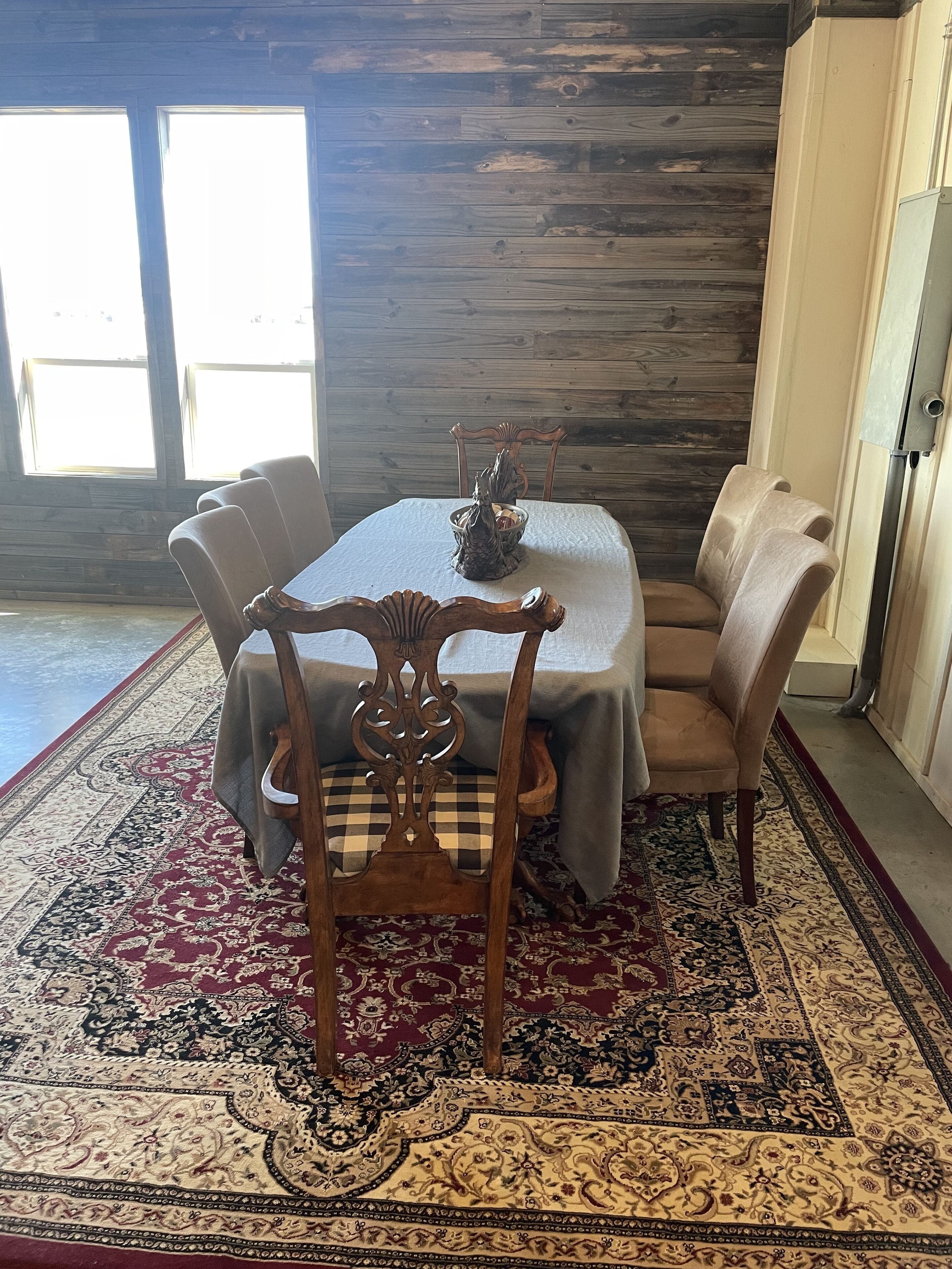 Comedor con una mesa dispuesta para seis personas, rodeada por seis sillas, un tapete estampado y una pared de madera.