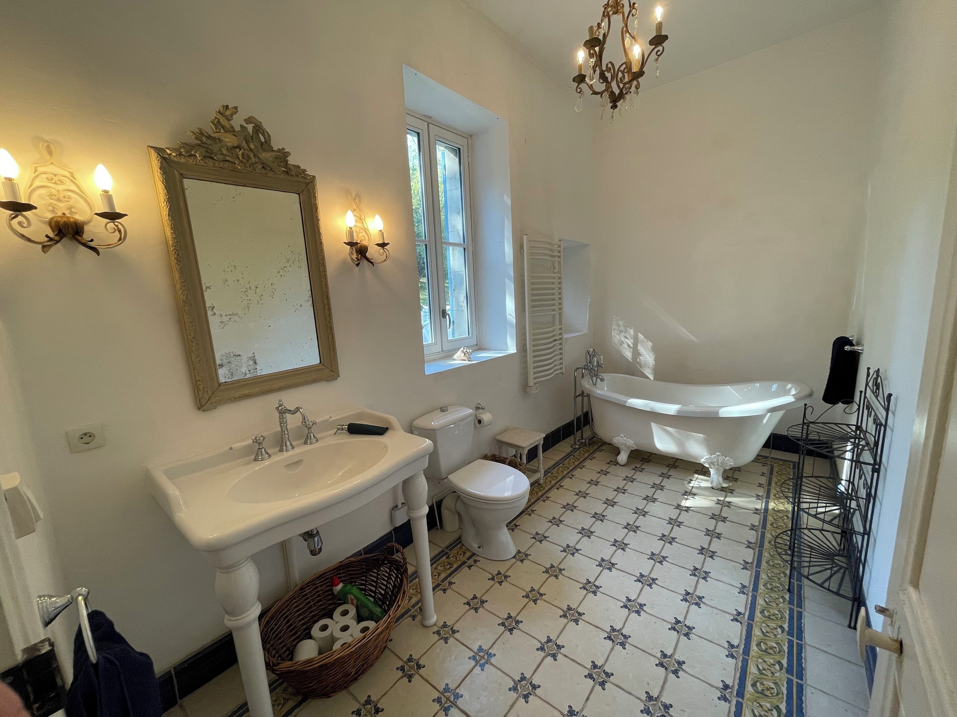 A bathroom with a sink, toilet, and bathtub. There is a window, a chandelier, and a mirror.