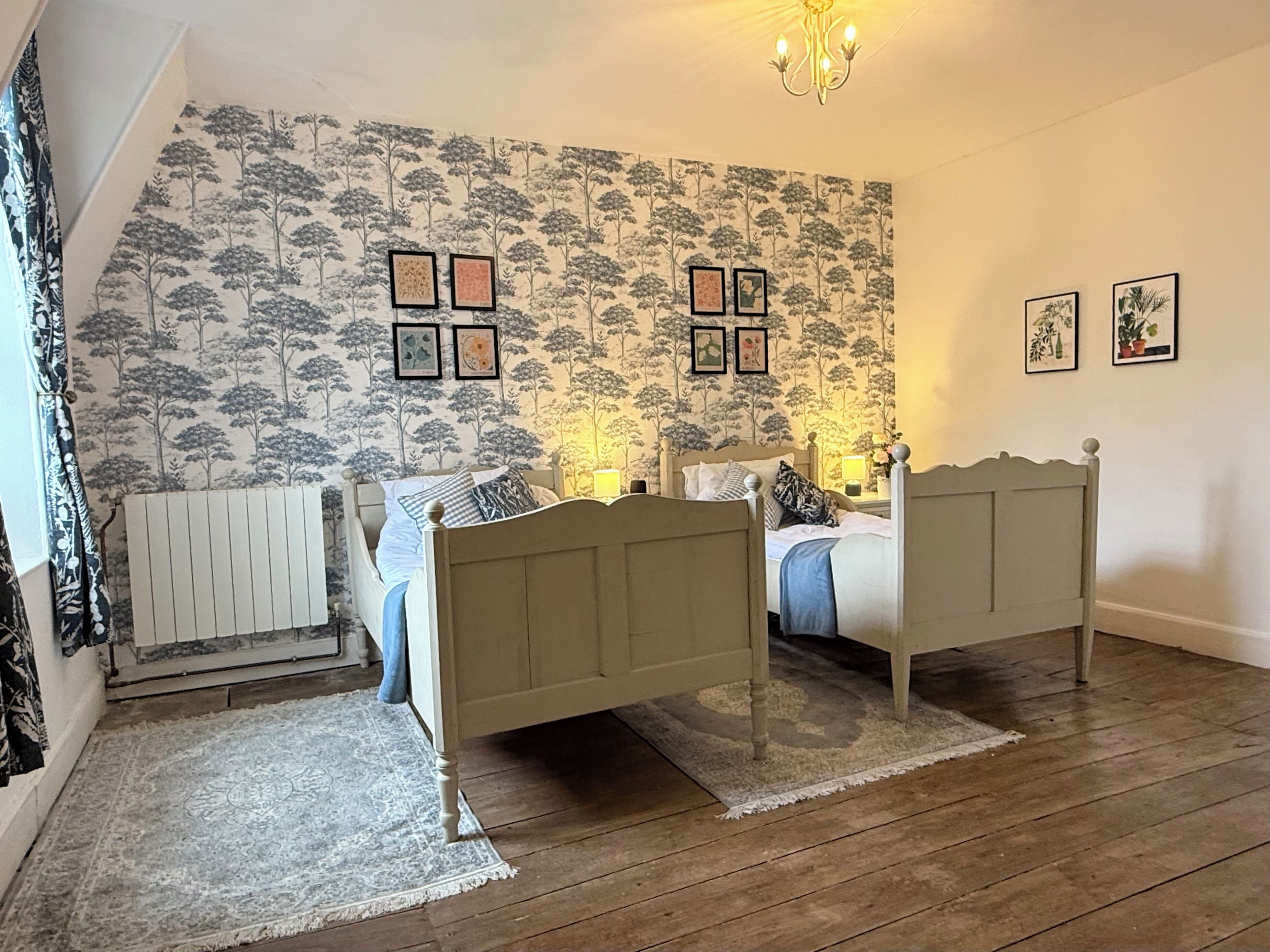 A bedroom with two single beds, a radiator, a patterned wallpaper, and framed pictures on the wall.