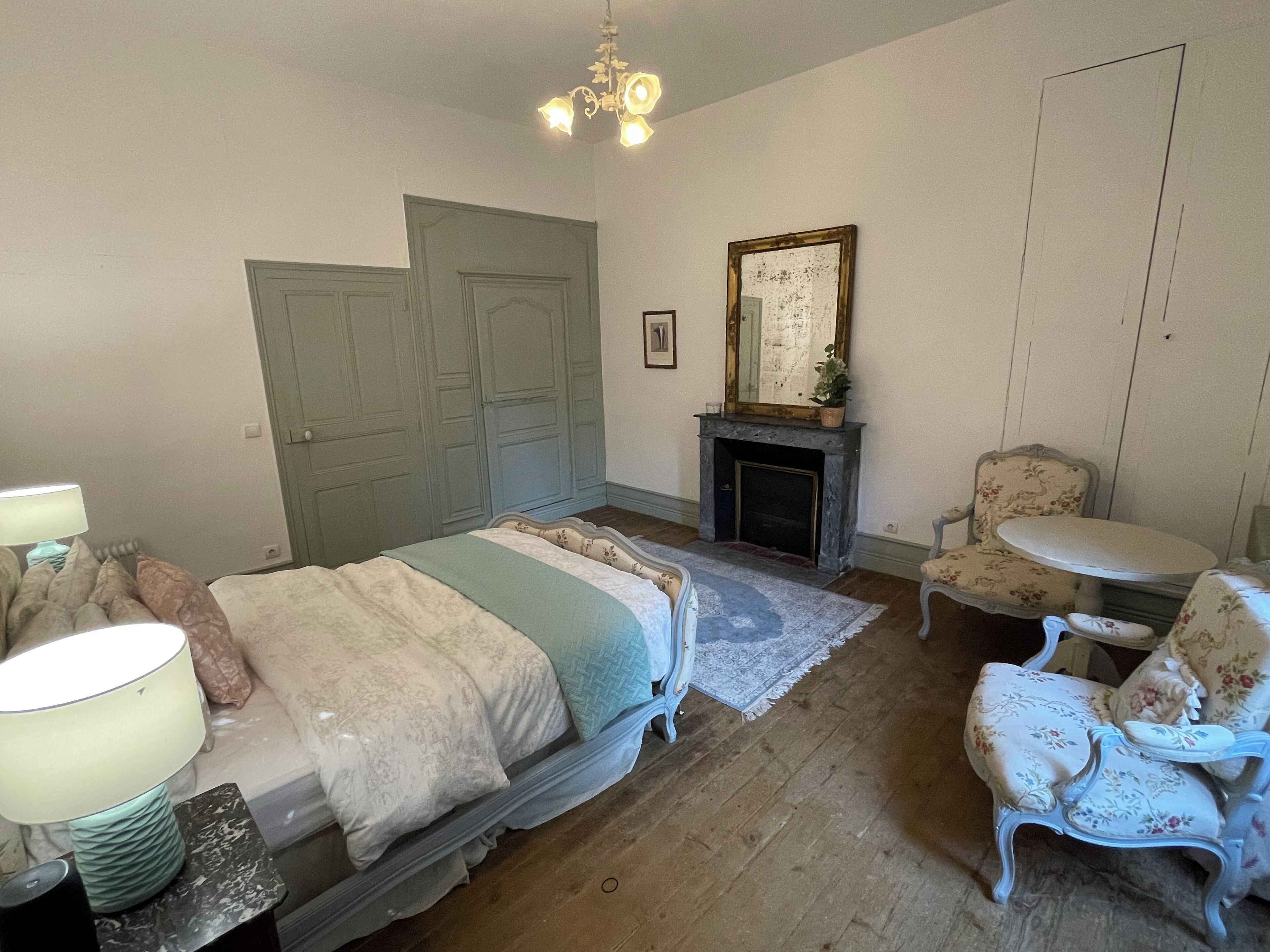A bedroom with a bed, a chair, a small table, a fireplace, and a chandelier.