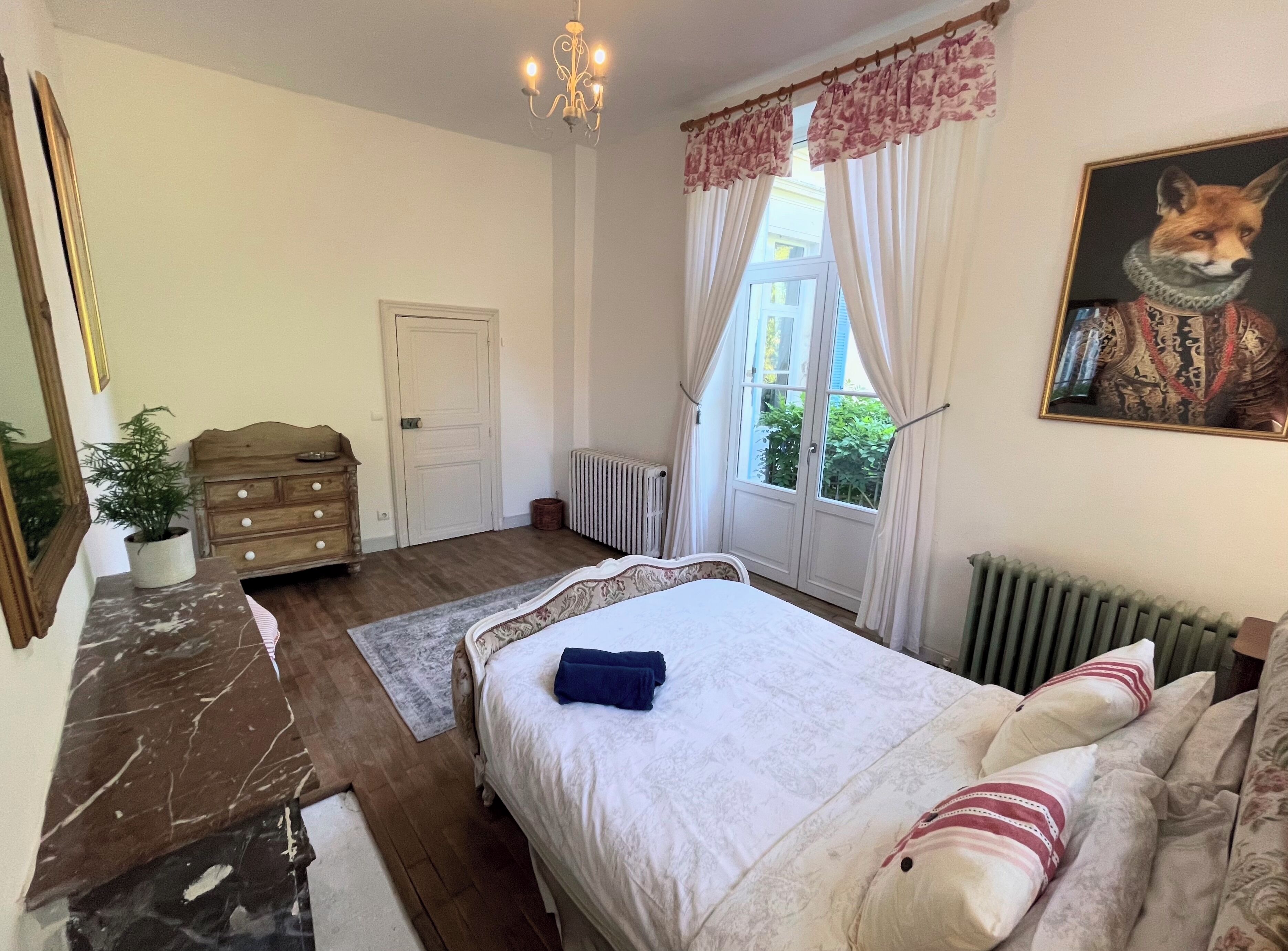 Een slaapkamer met een bed, een dressoir, een radiator, een raam met gordijnen en een schilderij van een vos.