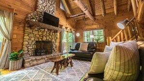 Un interior de cabaña acogedor, con chimenea de piedra, paredes de madera, un sofá de cuero y una mesa de comedor.