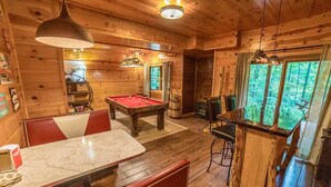 Interior de una cabaña de madera con mesa de billar, mesa de futbolín, una mesa de comedor y una zona de bar.