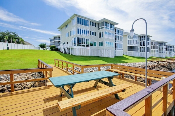 A multi-story building with a white exterior and large windows, a wooden deck with benches, and a clear sky.