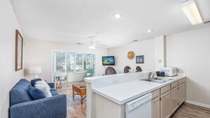 A modern kitchen with a central island, a dining area with a blue sofa, and a view of the outdoors through large windows.