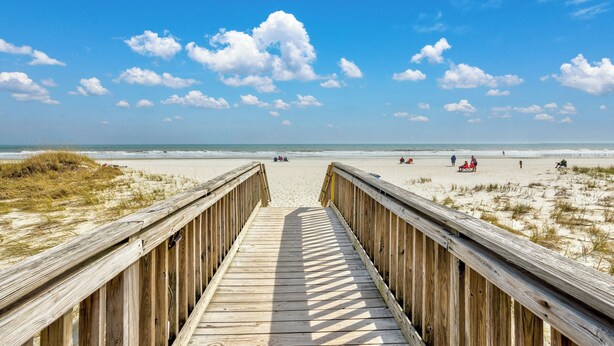 A wooden boardwalk leading to a sandy beach with a clear sky and scattered clouds.