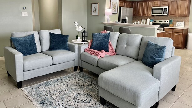 Un salon avec un canapé d’angle gris, deux tables d’appoint, un tapis à motifs et une cuisine en arrière-plan.