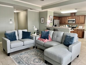 A living room with a grey sectional sofa, two side tables, a patterned rug, and a kitchen in the background. - Cozy Coastal Cottage that is 15-minute drive to the beach! (Corpus Christi)