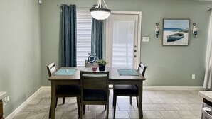Un coin repas avec une table et des chaises, un luminaire au plafond, une porte avec des stores et un tableau encadré au mur.