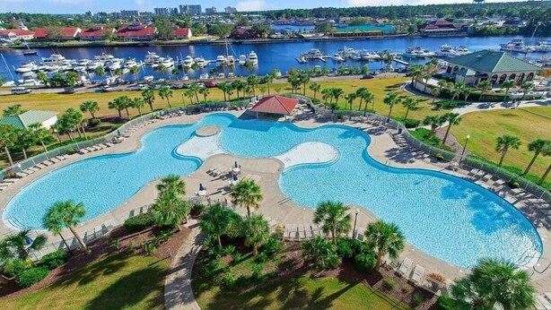A large, irregularly shaped swimming pool with a surrounding deck and palm trees, set against a backdrop of a marina with boats and buildings. - Intercostal Waterway Hotel Style Room -- 5 min from the beach! (North Myrtle Beach)