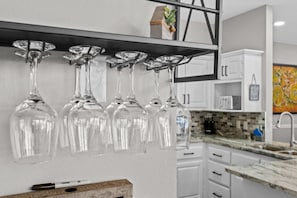 Une cuisine avec des meubles blancs, un plan de travail en granit et un porte-bouteilles à vin suspendu, muni de six verres à vin transparents.