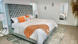 A bedroom with a tufted headboard, a bed with white and orange bedding, a bedside lamp, and a large mirror on the wall.