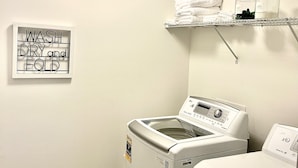 A laundry area with a washing machine, a dryer, a shelf with folded towels, and a sign that says "WASH DRY and FOLD".
