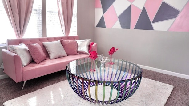 A pink sofa with white and pink cushions, a glass coffee table with a colorful striped base, and a geometric wall art piece.