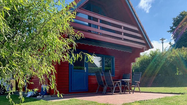 A red wooden house with a large window and a door, a patio with chairs, and a tree in the foreground. - 2 bedroom cozy home in Boiensdorf (Boiensdorf)