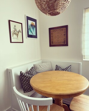 A dining area with a round table and patterned cushions on a bench, featuring framed artwork and a woven light fixture.