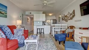 A living room with a red sofa, a white coffee table, and a blue ottoman. There is a kitchen area with white cabinets and a dining table with chairs.