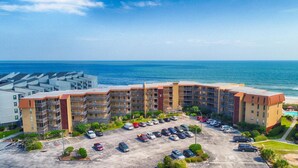 A multi-story hotel with a parking lot in front, located near a beach with the ocean in the background.