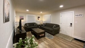 A living room with a sectional sofa, a coffee table, and a TV. There are two doors, one with a sign above it, and a wall-mounted picture.