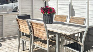 Un patio con una mesa y sillas de madera, una planta en maceta con flores rojas y un coche blanco estacionado cerca.