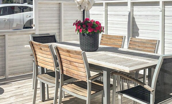 Eine Terrasse mit einem Holztisch und -stühlen, einem Blumentopf mit roten Blüten sowie ein in der Nähe geparktes weißes Auto.