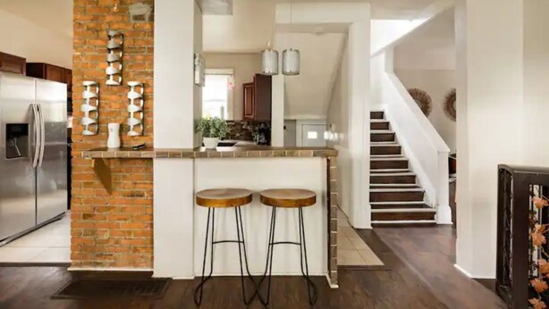 Une cuisine moderne avec un mur d’accent en briques, deux tabourets de bar, un réfrigérateur en acier inoxydable et un escalier menant à un autre niveau.