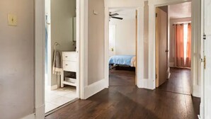 Un couloir au plancher de bois menant à une chambre à coucher équipée d’un lit et d’un ventilateur de plafond.