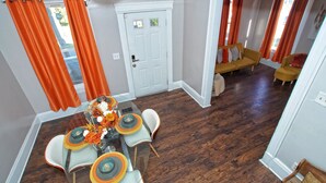 Un coin repas avec une table dressée pour quatre personnes, une porte blanche et un salon avec un canapé jaune et des rideaux orange.