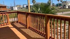 Una terraza de madera con techo metálico, rodeada de palmeras y casas al fondo.