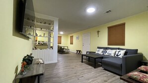 Una sala de estar con un sofá gris, una mesa de centro y un televisor montado en la pared. En el fondo, hay un comedor con una mesa y sillas.