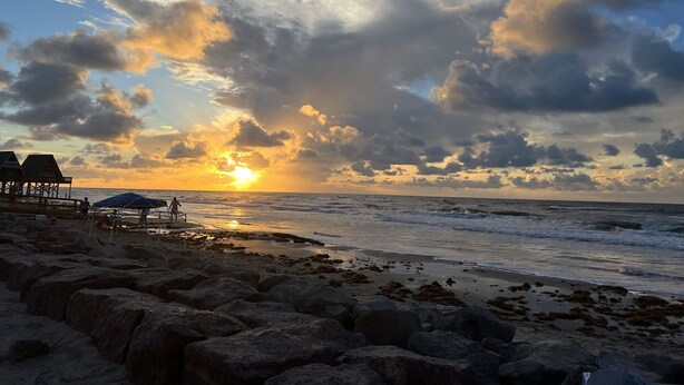 Una playa con un atardecer, costa rocosa y una estructura con sombrillas.