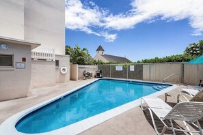 A rectangular swimming pool with a white border, surrounded by a fence, and a few lounge chairs. - Partial Ocean View Condo in Center of Kailua Kona (Kailua Kona)