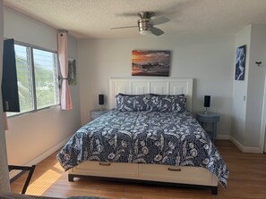 A bedroom with a bed, a ceiling fan, a window with curtains, a bedside table with a lamp, and artwork on the wall.