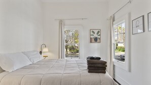 A bedroom with a bed, a nightstand, a window with curtains, and a view of the outside.