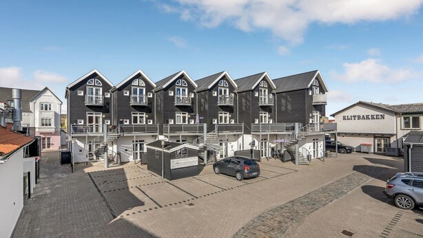 A row of dark-colored, multi-story buildings with balconies, a parking area, and a sign for 'KLITTEBAKKEN'.