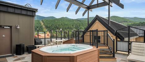 A hot tub on a patio with a view of mountains, a covered outdoor seating area, and a building with a metal exterior.