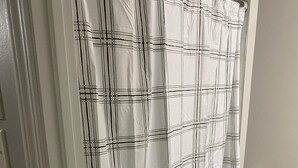 A bathroom with a white door, a white shower curtain with a plaid pattern, a black bath mat, and a toilet paper holder on the wall.