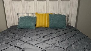 A bed with a grey quilted mattress, a white headboard with paneling, and three pillows in green, yellow, and plaid.