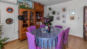 A dining area with a round table covered in a blue and purple textured tablecloth, surrounded by purple upholstered chairs. A wooden cabinet with glass doors is to the left, filled with various items.